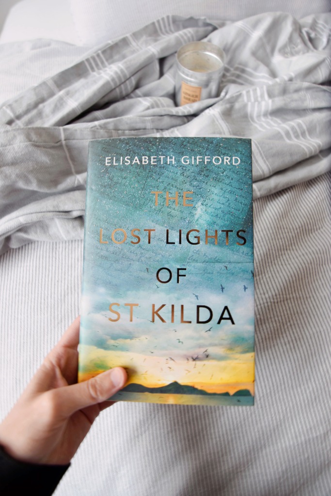 A hand holding a copy of The Lost Lights of St Kilda by Elisabeth Gifford against a backdrop of a bed with an unlit candle atop a blanket